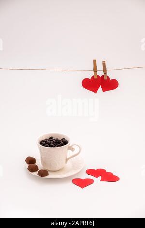 Tazza di caffè con biscotti al cioccolato a forma di cuore su sfondo bianco e steppe di vestiti e cuori di carta rossa sulla corda sopra. Concetto di giorno di San Valentino Foto Stock