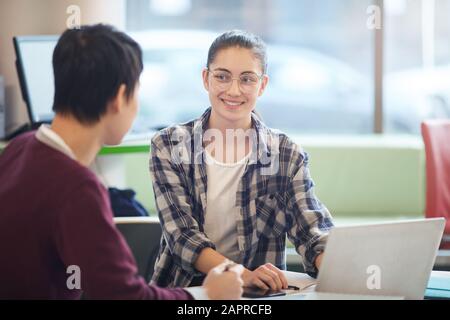 Giovane donna che usa un computer portatile e sorride alla sua collega mentre lavora al tavolo in ufficio Foto Stock