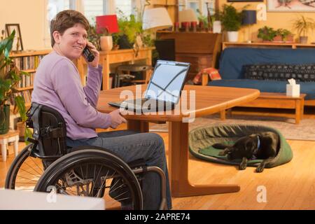Persone disabili in sedia a rotelle che utilizzano un computer portatile a casa e parlando al telefono con il suo cane nel sfondo Foto Stock