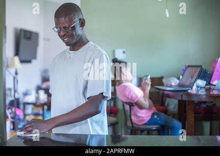 Una famiglia a casa, il padre che puliva contatori e la figlia in background utilizzando uno smartphone e un computer portatile Foto Stock
