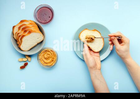 Vista dall'alto, piatto processo di cottura della colazione, spalmando il pane, toast con burro di arachidi, cremoso pasta di arachidi da donne mani su blu colorato Foto Stock