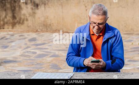 Uomo anziano che guarda e tocca lo schermo dello smartphone con il giornale sul tavolo mentre si siede nel parco Foto Stock