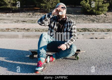 Giovane uomo moderno con un cappello di lana e occhiali da sole seduti sul longboard avendo un caffè Foto Stock