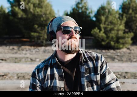 Giovane uomo moderno con occhiali da sole, cappello di lana e cuffie Foto Stock