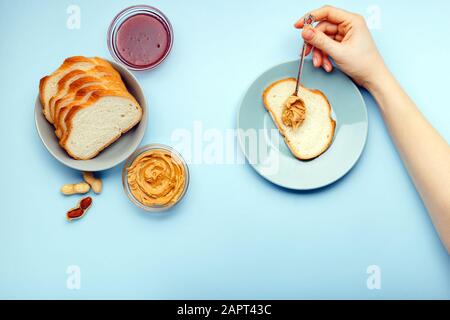 Vista dall'alto, piatto processo di cottura della colazione, spalmando il pane, toast con burro di arachidi, cremoso pasta di arachidi da donne mani su blu colorato Foto Stock