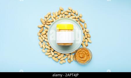 Pasta di arachidi cremosa in vaso di vetro con tappo giallo con mock-up e burro di arachidi in una lastra di vetro. Arachidi nella buccia sparse su sfondo blu Foto Stock
