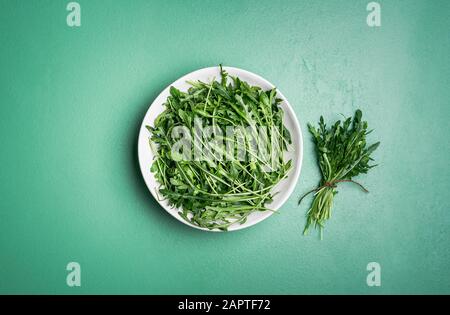 Mazzo di rucola fresca in un piatto bianco e un mazzo di rucola vicino ad esso su un tavolo verde alla menta. Sopra vista di rucola raccolta. Preparazione dell'insalata di rucola. Foto Stock