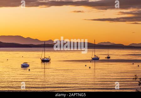 Barche a vela ormeggiate al tramonto con la luce del sole dorata che si riflette l'acqua tranquilla e una costa silhouette Foto Stock