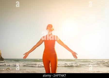 donna che indossa abbigliamento sportivo arancione in piedi dal mare con le braccia fuori e faccia fino al sole Foto Stock