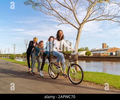 Quattro adolescenti che si divertono all'aperto, accanto a un fiume, con pattini a rotelle e una bicicletta Foto Stock