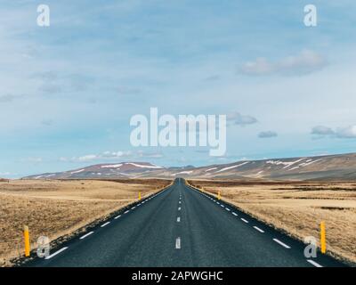 Strada circondata da colline coperte di verde e neve sotto Un cielo nuvoloso in Islanda Foto Stock