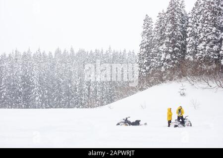 Due uomini in giacche gialle con motoslitte (macchina da neve, slitta a motore, slitta a motore, skimobile, motoslitta, slitta o Ski-Doo) parlano sulla neve. Foto Stock