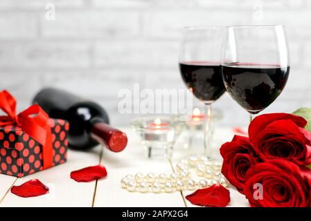 Concetto di San Valentino. Due bicchieri di vino, rose rosse, regalo, filo di perle, petali di rosa, bottiglia di vino e candele accese su un tavolo di legno bianco. Foto Stock