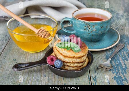 Colazione a base di pancake in padelle in ghisa, frutti di bosco freschi, miele e tè nero in stile rustico Foto Stock