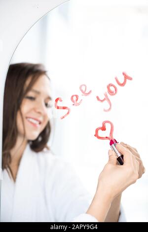 Mano di felice scrittura femminile vedere e disegnare il cuore con rossetto sullo specchio Foto Stock