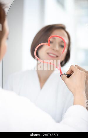 Riflesso di giovane donna flirty che disegna il cuore con rossetto cremisi sullo specchio Foto Stock