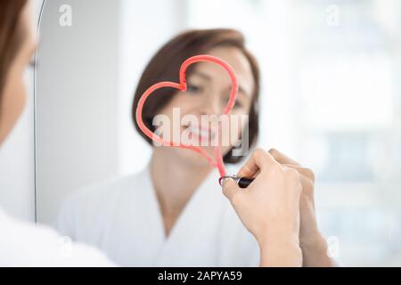 Riflesso di giovane brunetta femminile in accappatoio bianco disegno cuore sullo specchio Foto Stock