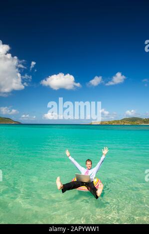 Uomo d'affari che lancia le braccia e le gambe in festa mentre galleggia in una baia tropicale con il suo portatile Foto Stock