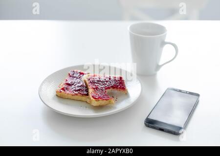 La colazione è a tavola. Una tazza di caffè e toast con marmellata di ciliegie accanto al telefono. Foto Stock