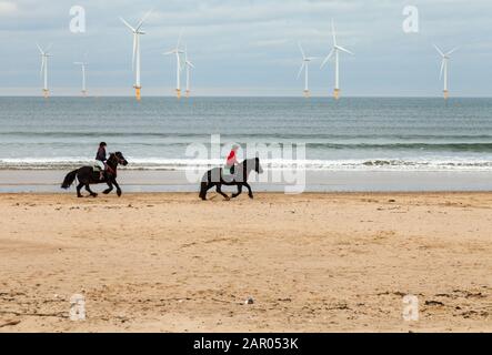 Due donne equitazione cavalli sulla spiaggia Redcar con le turbine eoliche in background nel nord est Inghilterra, Regno Unito Foto Stock