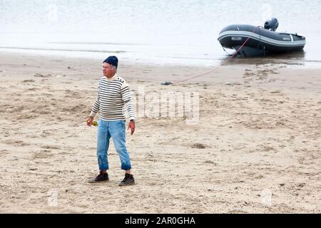 Bevuto marinaio con bottiglia di vino in mano sta camminando sulla spiaggia, con la sua barca sullo sfondo Foto Stock