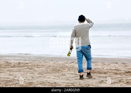 Marinaio con bottiglia di vino in mano si erge sulla spiaggia e si affaccia sul mare Foto Stock