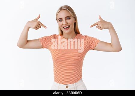 Orgogliosa, boastful bella ragazza elegante con capelli biondi, tatuaggi, in piedi a righe t-shirt, che si punta come bragging, sassy sorridente, spettacolo Foto Stock