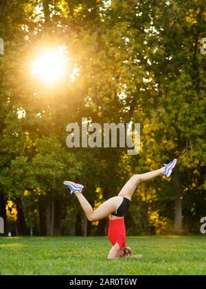 Una giovane ragazza pratica yoga in natura. Donna esegue un headstand in un parco. Concetto - stile di vita sano e attivo Foto Stock