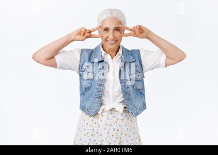 Allegro, felice sorridente vecchia donna moderna, granny che mostra la pace o la vittoria segni vicino agli occhi, insegnando alla generazione più giovane come danza stile discoteca, sorridente Foto Stock