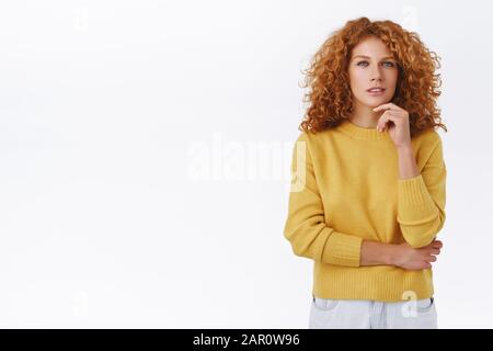 Donna curly meditata e dall'aspetto serio nel maglione giallo hanno un pensiero interessante, tocca il mento e l'unzione come vedere qualcosa di interessante Foto Stock