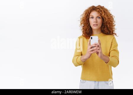 Tenera, affascinante cute redhead donna con capelli ricci in maglione giallo, tenere smartphone, prendere selfie in specchio, fotografare qualcosa di stupefacente e. Foto Stock