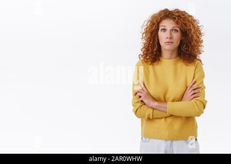 Scettico attraente donna curly redhead in maglione giallo guardare con dubbio e giudizio alla fotocamera, croce braccia petto chiuso posa, ascoltare qualche strano Foto Stock