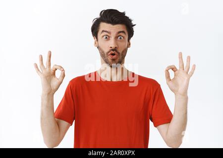 Impressionato, emozionato buon-osservare l'uomo caucasico con la barba nella t-shirt rossa, mostrando okay, gesto ok, giudicante qualcosa impressionante, dica wow, tasso meraviglioso Foto Stock