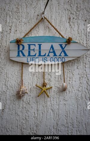 Cartello in legno a forma di barca che dice Relax, la vita è buona. Concetto di pensiero positivo, appeso su muro bianco con tre conchiglie di mare su archi Foto Stock