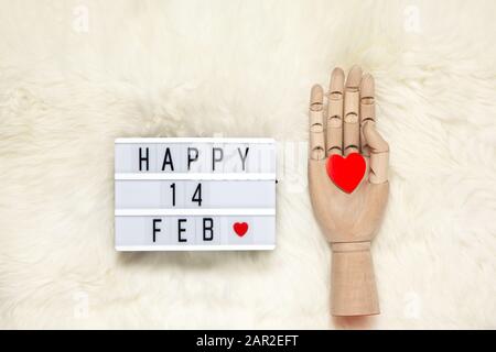 Il modello di mano umana di legno alla moda tiene il cuore rosso, la scatola luminosa con l'iscrizione Felice 14 FEBBRAIO che significa giorno di San Valentino su tappeto di pelliccia bianca. Disposizione piatta. Febbraio Foto Stock