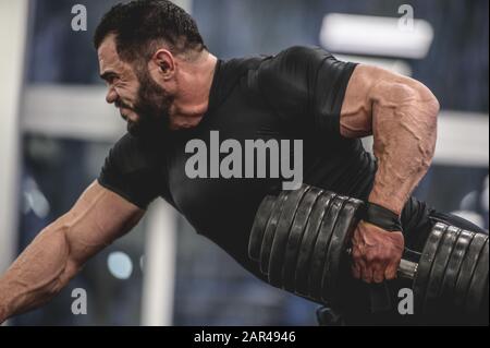 forte giovane bearded caucasico in nero sportswear tirando pesi pesanti manubrio con una mano di allenamento schiena e braccia in palestra sport allenamento Foto Stock