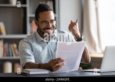 Felice soddisfatto maschio africano professionista leggere buone notizie nel documento Foto Stock