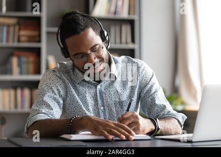 Sorridente studente africano maschio professionista indossare cuffia e-learning prendere appunti Foto Stock