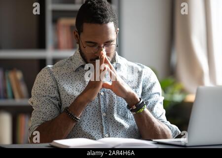 Giovane uomo d'affari americano africano che prega sedersi al banco di lavoro Foto Stock