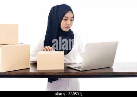 Asian Le donne musulmane merchant lavora con il computer portatile da casa su un tavolo di legno con pacco postale le scatole di cartone la preparazione per la spedizione del suo ordine Foto Stock