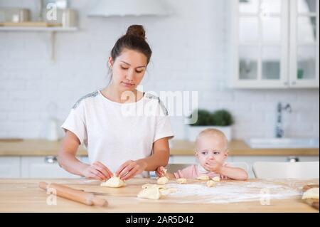 la madre sculpts torte, stando in piedi al piano di appoggio in una cucina luminosa, e una piccola figlia nelle vicinanze mangia pasta Foto Stock