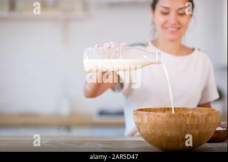 ragazza che versa il latte da una bottiglia di plastica in una ciotola di legno, sorridendo sullo sfondo. Foto Stock