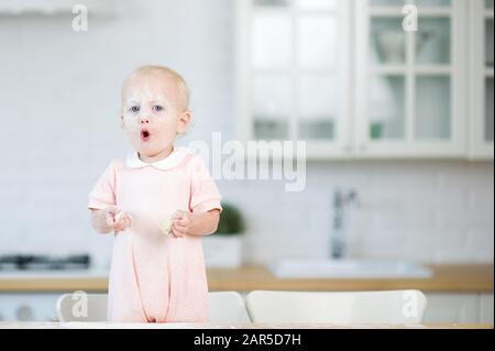 baby ragazza con farina sul viso e fette di pasta nelle mani si alza dietro il piano di lavoro della cucina e guarda nella cornice, aprendo la bocca Foto Stock