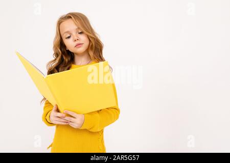 Felice adolescente ragazza con i capelli rossi, felpa con cappuccio e pantaloni gialli letto grande libro giallo isolato su sfondo bianco Foto Stock