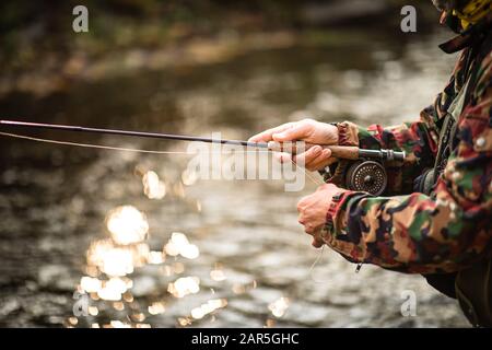 Vista ravvicinata delle mani di un pescatore a mosca che lavorano la linea e la canna da pesca mentre la pesca con la mosca su di uno splendido fiume di montagna per la trota iridea Foto Stock