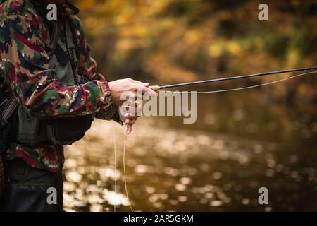 Vista ravvicinata delle mani di un pescatore a mosca che lavorano la linea e la canna da pesca mentre la pesca con la mosca su di uno splendido fiume di montagna per la trota iridea Foto Stock