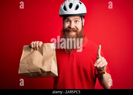 Red head irlandese consegna uomo con la barba indossare il casco bici e tenere takeaway sacchetto di carta sorpreso con un'idea o domanda dito di puntamento con felice Foto Stock