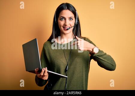 Giovane operatore donna con occhi blu che lavora sul call center utilizzando computer portatile e cuffia con un dito a sorpresa rivolto verso se stesso Foto Stock