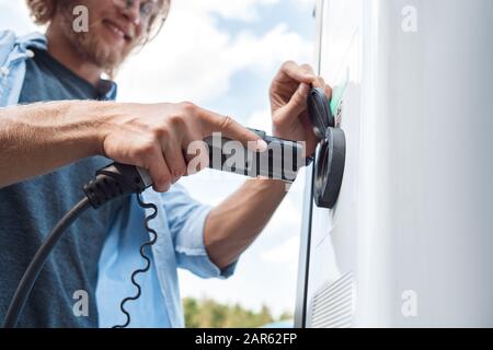 Trasporto. Giovane uomo che viaggia in auto elettrica sosta presso la stazione di ricarica collegare il caricabatterie da vicino sorridendo allegro Foto Stock