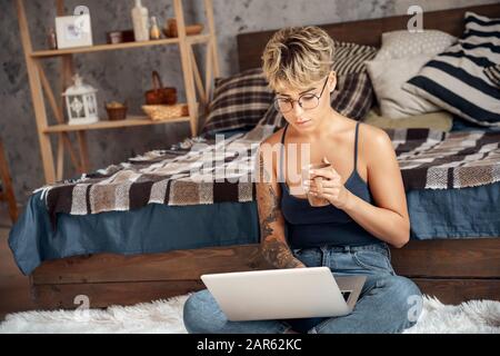 Side Hustle. Giovane donna capelli corti in occhiali seduti sul pavimento lavorando online su laptop caffè da bere pensive Foto Stock